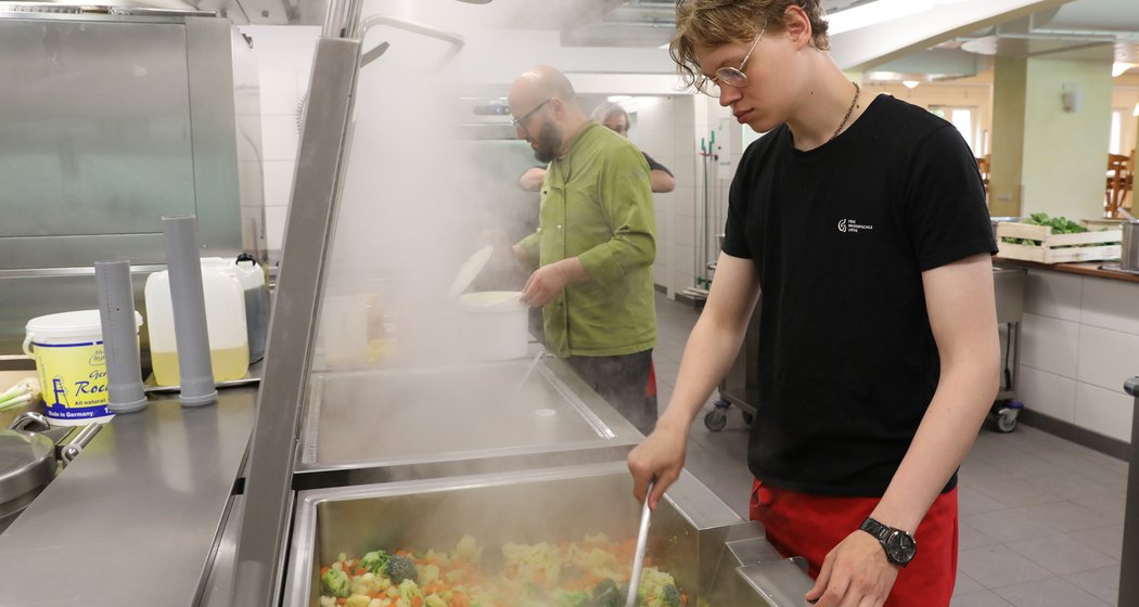 In einer Großküche arbeiten zwei Personen. Eine rührt mit einem großen Löffel in einer dampfenden Pfanne mit gemischtem Gemüse um, während eine andere im Hintergrund Speisen zubereitet. Die Küche ist sauber und gut beleuchtet.