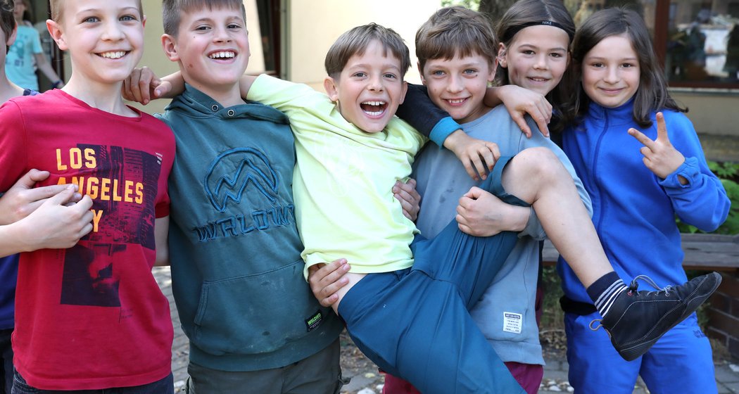 Sechs lächelnde Kinder posieren im Freien; vier von ihnen halten einen lachenden Freund in blauen Shorts und einem gelben T-Shirt in die Höhe, während ein weiteres neben ihnen steht und das Peace-Zeichen macht. Sie wirken an diesem sonnigen Tag fröhlich und ausgelassen.