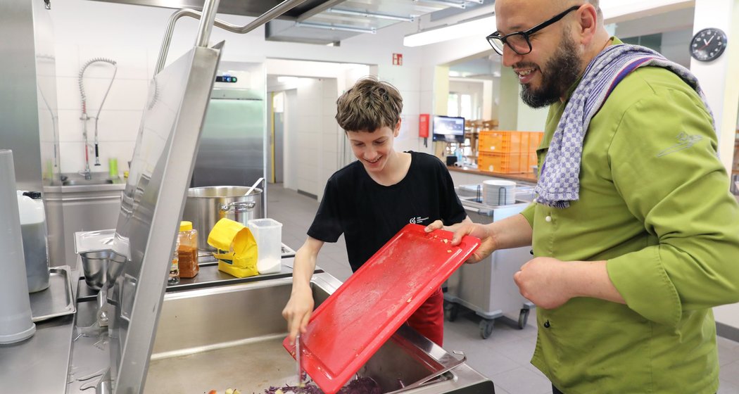 Ein Koch und ein junger Mensch bereiten gemeinsam in einer Großküche Essen zu und lächeln, während sie gehackte Zutaten in einen großen Topf geben.