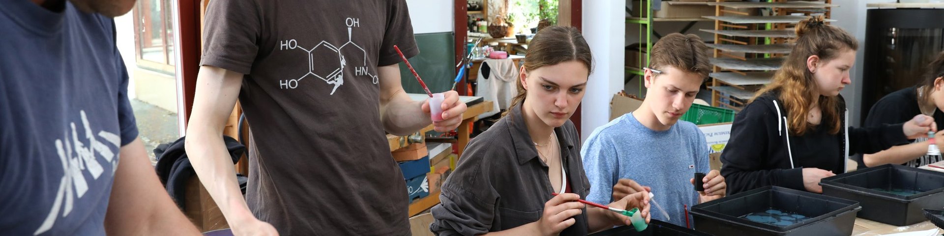 Mehrere Jugendliche und ein Erwachsener sitzen um Tische herum und sind mit einer künstlerischen Aktivität beschäftigt, bei der Farbe und Wasserschalen zum Einsatz kommen – in einem hellen, geräumigen Raum, der mit Malutensilien und Materialien gefüllt ist.