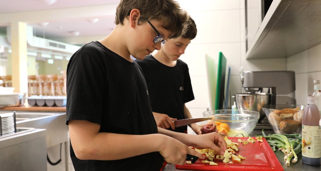 Zwei Jungen in schwarzen Hemden schneiden in einer Küche Gemüse auf einem roten Schneidebrett. Um sie herum sind Schüsseln, Küchenutensilien und Zutaten zu sehen, während sie arbeiten.