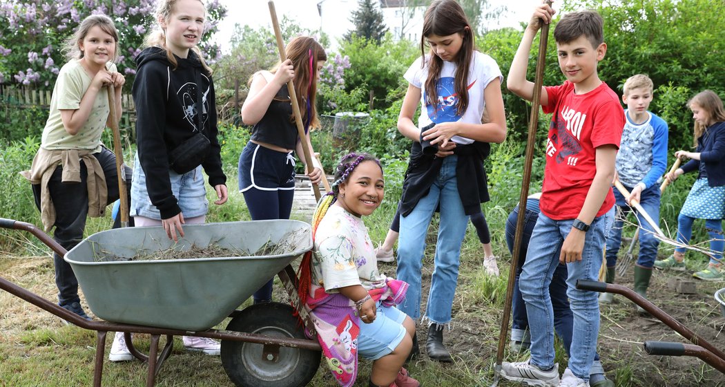 Eine Gruppe von Kindern arbeitet gemeinsam in einem Garten; einige halten Werkzeuge und eine Schubkarre in der Hand. Sie lächeln und scheinen Spaß an der Aktivität im Freien zu haben. Im Hintergrund sind Grünflächen und ein Gebäude zu sehen.