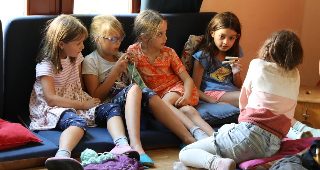 Fünf junge Mädchen sitzen eng beieinander auf einem Sofa und auf dem Boden, unterhalten sich und sind mit verschiedenen Aktivitäten beschäftigt – in einem gemütlichen Raum mit Kissen und sanftem Licht, das durch die Fenster hereinströmt.