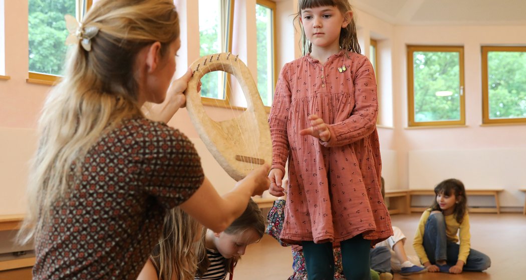 Eine Frau hält einem kleinen Mädchen in einem rosa Kleid, das in einem Klassenzimmer danach greift, eine hölzerne Leier hin. Im Hintergrund sitzen weitere Kinder auf dem Boden.