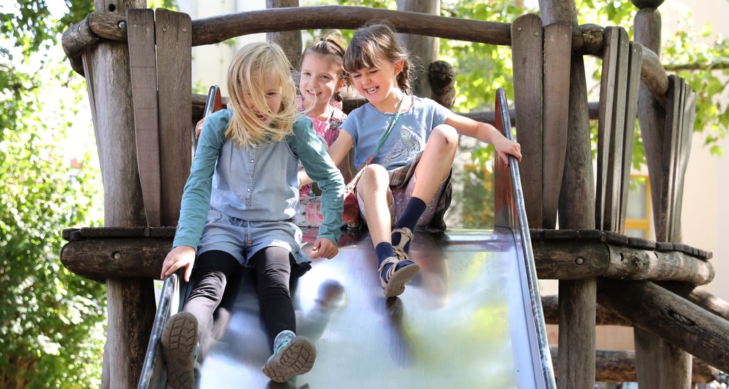 Drei kleine Kinder lächeln und rutschen an einem sonnigen Tag eine glänzende Metallrutsche hinunter, umgeben von Holzkonstruktionen und grünen Bäumen.