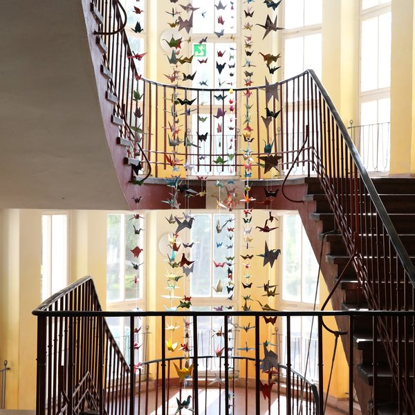 Ein Treppenhaus mit großen Fenstern und Papierkranichen, die in vertikalen Reihen von der Decke hängen und in dem sonnendurchfluteten Raum eine farbenfrohe, zarte Dekoration bilden.