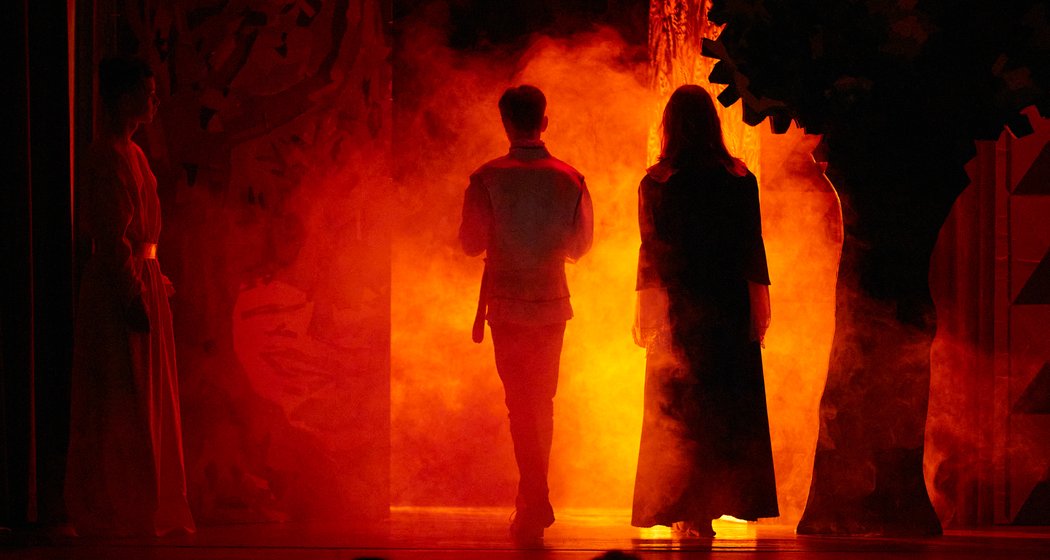 Drei Personen stehen auf der Bühne vor einem leuchtend feurig-orangefarbenen Hintergrund mit Rauch. Zwei stehen in der Mitte mit dem Rücken zur Kamera, eine steht links im Schatten. Die Szene vermittelt den Eindruck eines dramatischen oder theatralischen Moments.
