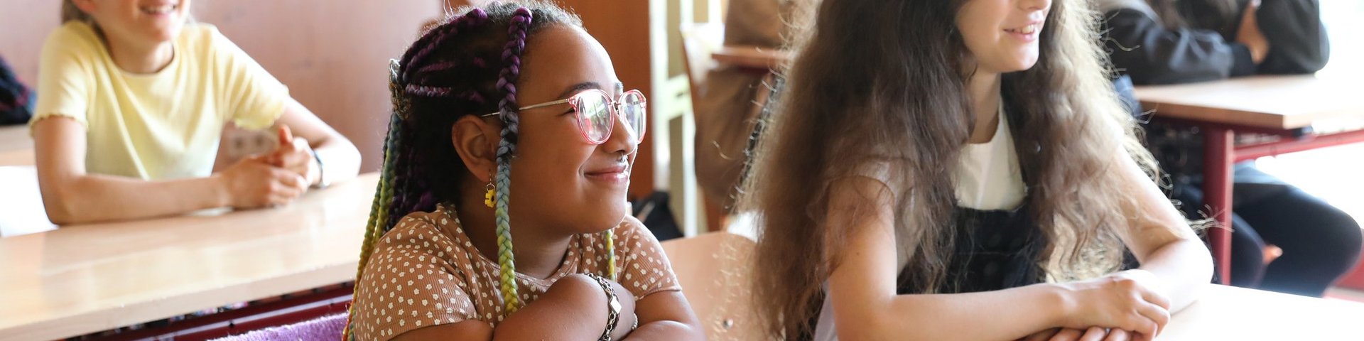In einem hellen Klassenzimmer sitzen mehrere Kinder an ihren Schreibtischen, lächeln und blicken nach vorne; sie wirken aufmerksam und glücklich.