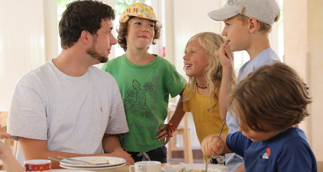 Ein Mann und vier Kinder sitzen um einen Tisch herum, lächeln und unterhalten sich beim Essen. Ein Kind steht dicht neben dem Mann, ein anderes konzentriert sich ganz auf sein Essen. Die Atmosphäre wirkt freundlich und ungezwungen.