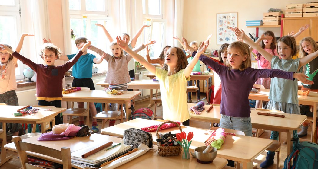 Eine Gruppe von Kindern steht mit erhobenen Armen neben den Schreibtischen in einem hellen Klassenzimmer und nimmt an einer interaktiven Aktivität teil. Schulmaterial, Taschen und Sonnenlicht erfüllen den fröhlichen Raum.
