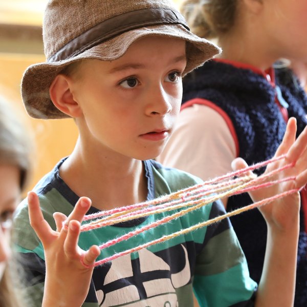 Ein kleiner Junge mit einer Mütze spielt mit einem bunten Faden und formt damit Muster zwischen seinen Fingern. In der Nähe sind weitere Kinder zu sehen, von denen eines im Hintergrund unscharf abgebildet ist. Die Szene scheint in einem Innenraum stattzufinden, möglicherweise in einem Klassenzimmer.