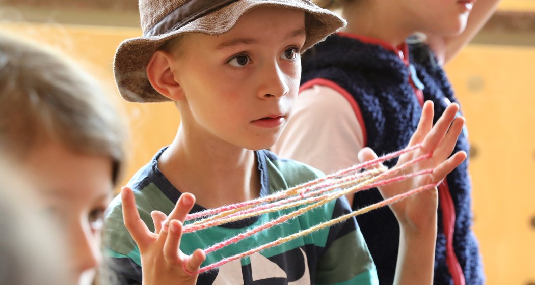Ein kleiner Junge mit einer Mütze spielt mit einem bunten Faden und formt damit Muster zwischen seinen Fingern. In der Nähe sind weitere Kinder zu sehen, von denen eines im Hintergrund unscharf abgebildet ist. Die Szene scheint in einem Innenraum stattzufinden, möglicherweise in einem Klassenzimmer.