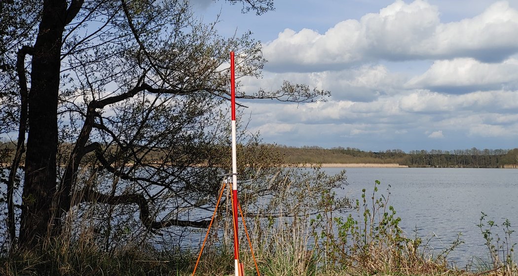 Ein rot-weißes Vermessungsstativ steht an einem See, umgeben von Gras und Bäumen, im Hintergrund sind Wolken und Bäume in der Ferne zu sehen.