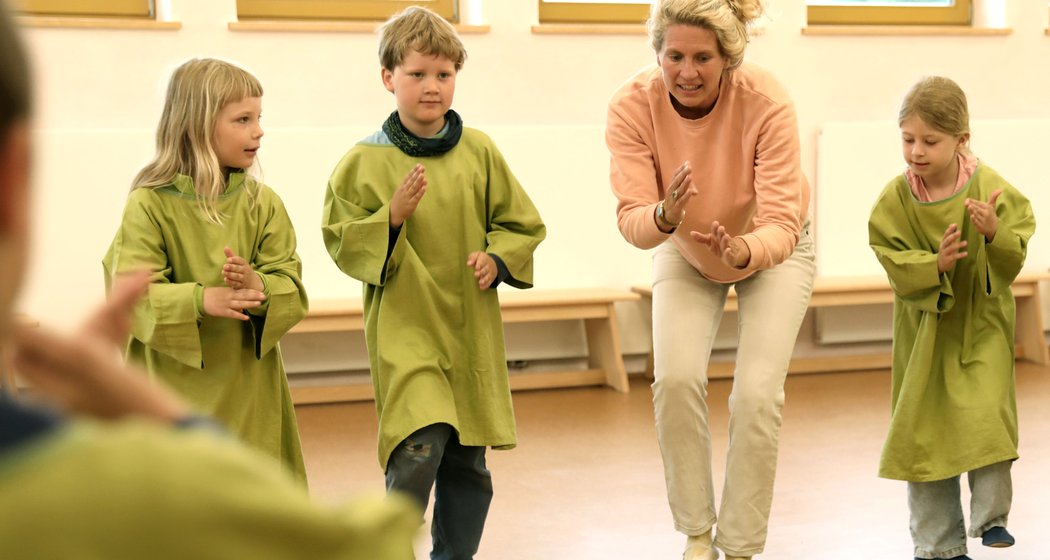 Eine Frau und drei kleine Kinder in grünen Kitteln stehen in einem Raum und ahmen ihre Hand- und Körperbewegungen nach, während sie an einer Gruppenaktivität oder einem Spiel teilnehmen.
