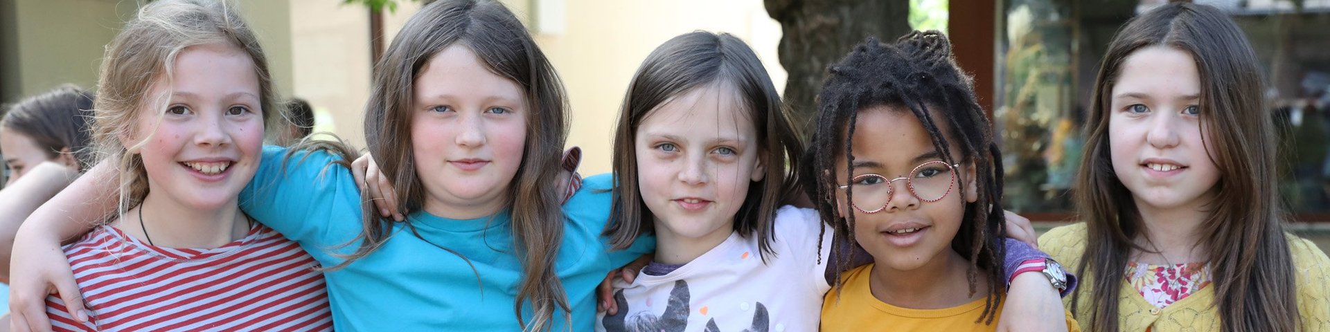 Fünf Kinder stehen draußen dicht beieinander und lächeln in die Kamera. Sie haben ihre Arme umeinander gelegt und tragen bunte Freizeitkleidung. Im Hintergrund sind Bäume und andere Menschen zu sehen.