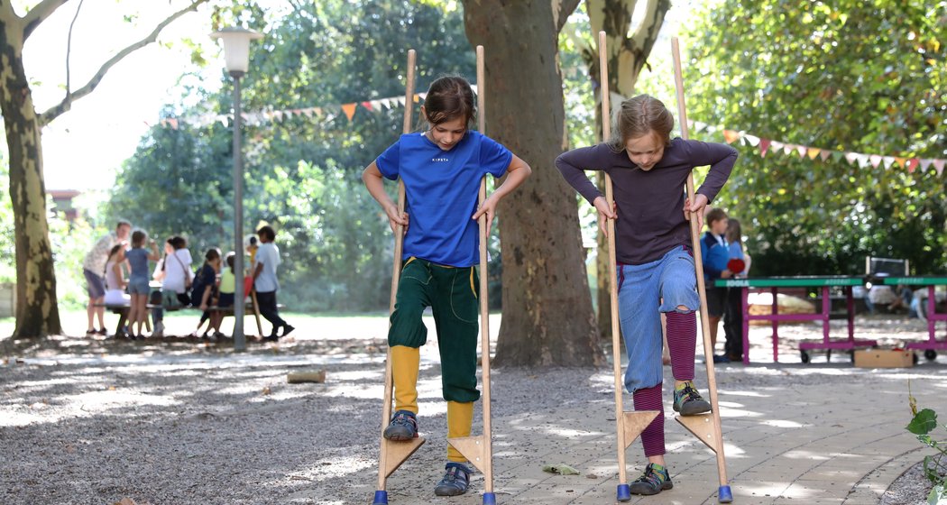 Zwei Kinder laufen in einem Park auf Holzstelzen und konzentrieren sich darauf, das Gleichgewicht zu halten. Im Hintergrund sind zwischen Bäumen und Wimpeln weitere Kinder und Erwachsene zu sehen, was auf eine fröhliche Veranstaltung im Freien hindeutet.