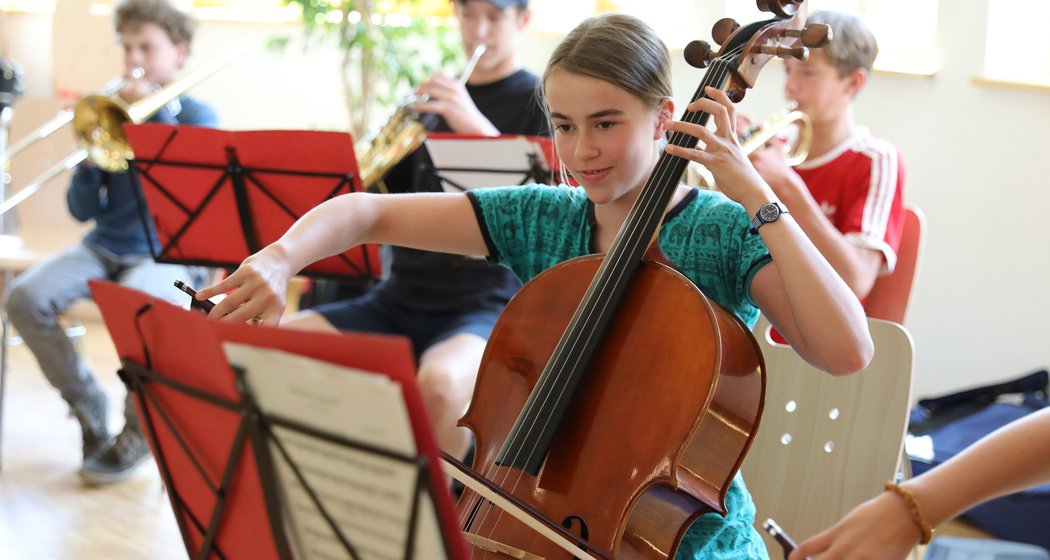 Ein Mädchen spielt Cello in einer Gruppe junger Musiker, zu der auch Blechbläser gehören, während einer Probe in einem hellen Raum, in dem Noten auf Notenständern liegen.