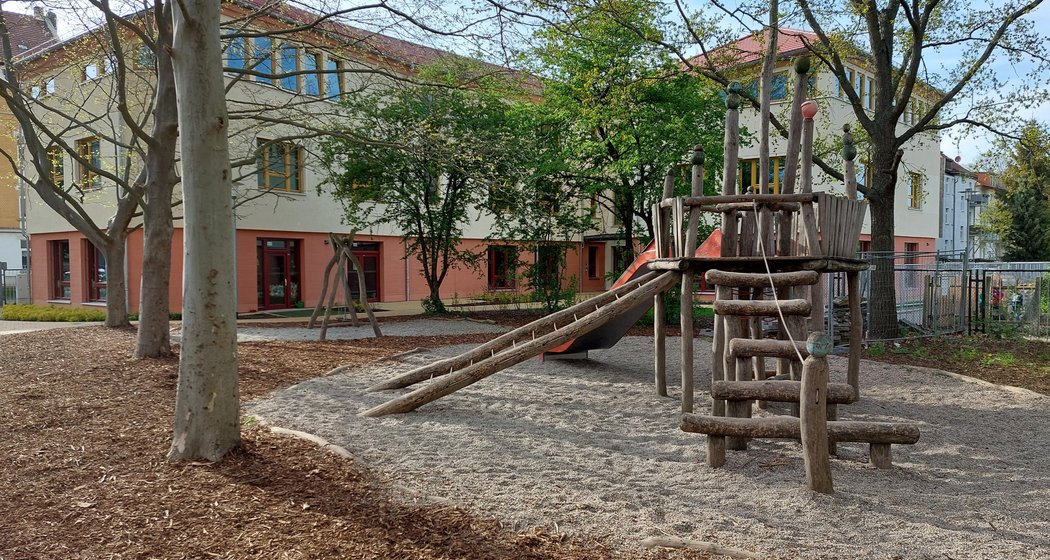 An einem sonnigen Tag liegt vor einem beigen Gebäude mit großen Fenstern ein Spielplatz mit einer Kletteranlage aus Holz und einer Rutsche, umgeben von Bäumen und Holzspänen.