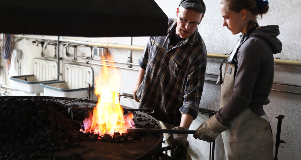 In einer Schmiede arbeiten zwei Personen. Ein Mann gibt einer Frau Anweisungen, während sie Metall über eine leuchtend orangefarbene Flamme hält; beide tragen in der Industriehalle Schutzkleidung.