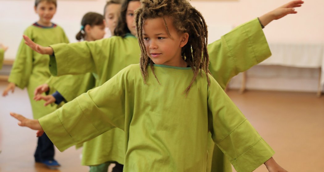 Kinder in weiten grünen Hemden stehen mit ausgestreckten Armen in einer Reihe in einem Raum und nehmen an einer Gruppenaktivität oder einem Tanz teil. Im Mittelpunkt steht ein Kind mit langen, lockigen Haaren, das ganz vorne in der Gruppe steht.
