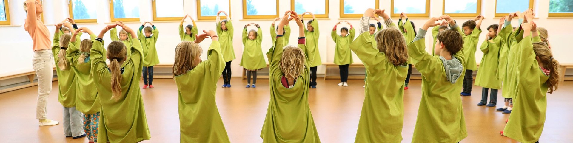 Eine Gruppe von Kindern in grünen Kitteln steht mit erhobenen Armen im Kreis und ahmt die Haltung eines Erwachsenen nach – in einem hellen Klassenzimmer mit großen Fenstern.