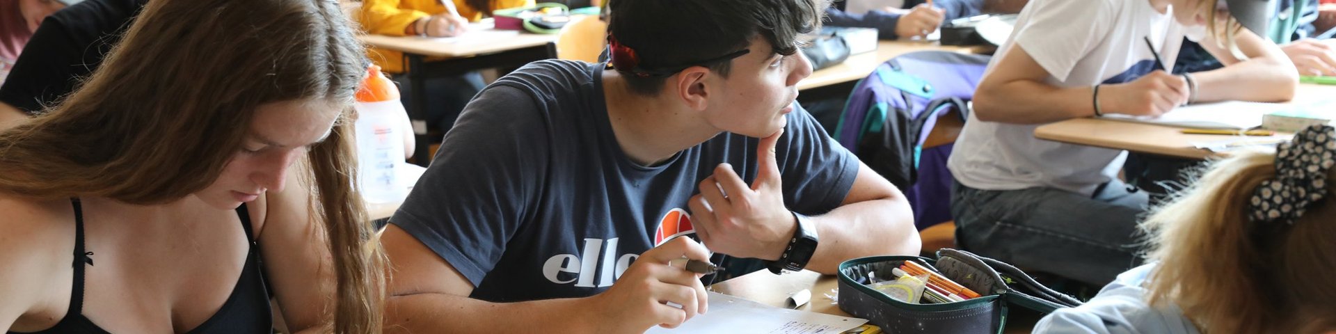Schüler, die an ihren Schreibtischen in einem Klassenzimmer sitzen, konzentrieren sich auf das Schreiben oder Lesen. Die meisten scheinen Teenager zu sein; auf ihren Schreibtischen liegen Bücher und Federmäppchen. Das Klassenzimmer hat rote Vorhänge, und durch die Fenster fällt Licht herein.