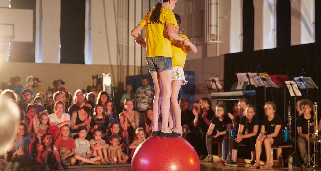 Zwei Mädchen in gelben T-Shirts balancieren in einer Turnhalle auf einem großen roten Ball und treten vor einem sitzenden Publikum aus Kindern und Erwachsenen auf. Im Hintergrund sind Musikinstrumente und Notenständer zu sehen.
