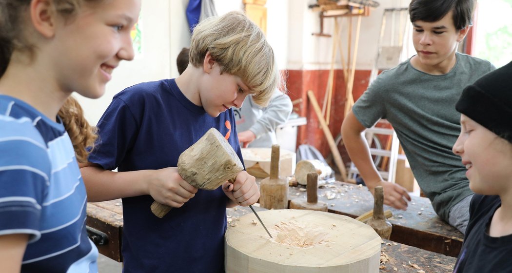 Vier Kinder arbeiten gemeinsam in einer Holzwerkstatt; ein Junge schnitzt mit Hammer und Meißel ein rundes Stück Holz, während die anderen zuschauen und lächeln. Auf dem Tisch liegen Werkzeuge und Holzspäne.