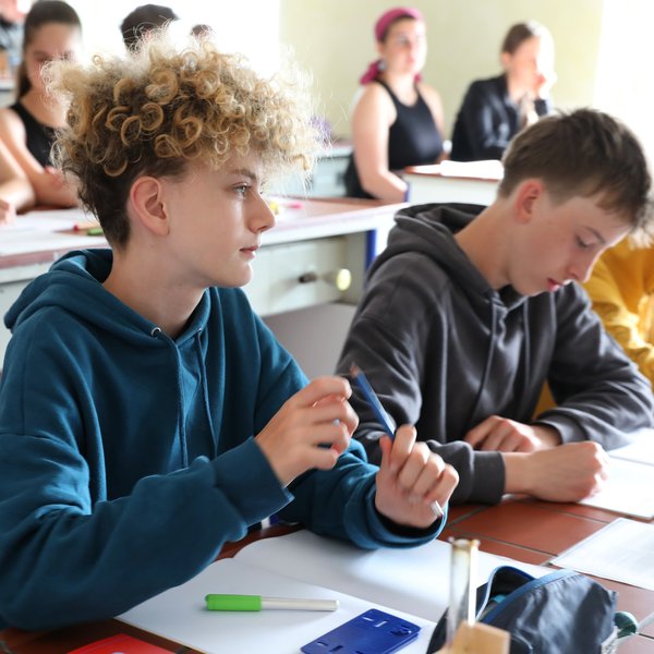 Mehrere Schüler sitzen an ihren Schreibtischen in einem Klassenzimmer und sind ganz in ihre Arbeit vertieft. Ein Schüler mit lockigem Haar und einem blauen Kapuzenpulli hält einen Stift in der Hand, während andere schreiben oder in ihre Unterlagen schauen. Auf den Schreibtischen liegen Schulutensilien verstreut.