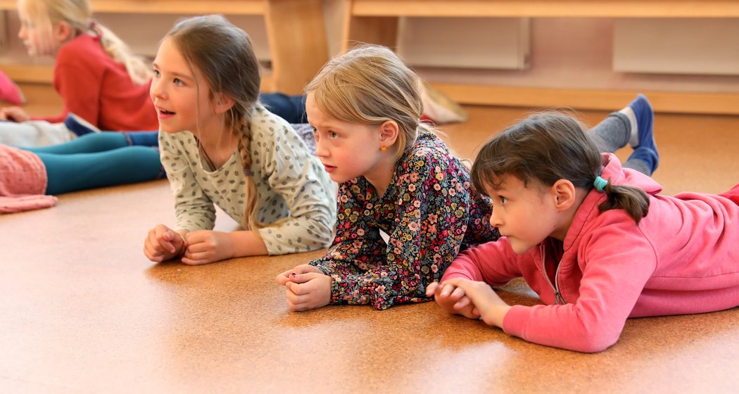 Drei kleine Kinder liegen drinnen auf dem Boden, stützen sich auf ihre Ellbogen und beobachten interessiert etwas. Im Hintergrund sind zwei weitere Kinder zu sehen. Alle tragen Freizeitkleidung.