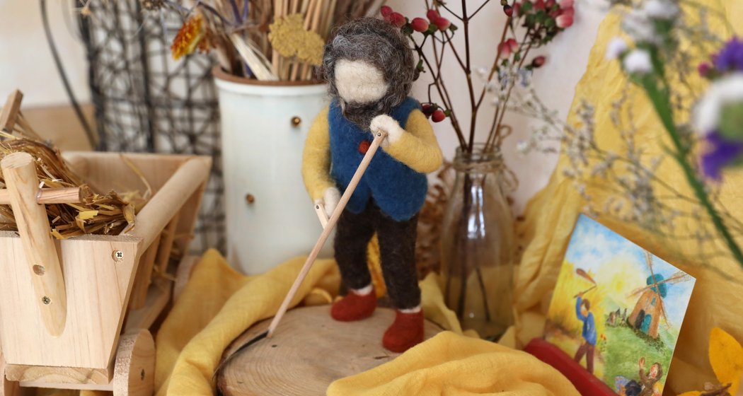 Eine Figur aus gefilzter Wolle gleitet an einer hölzernen Schubkarre, an getrockneten Blumen in Vasen und an einem kleinen Gemälde auf einer mit gelbem Stoff bezogenen Fläche vorbei.