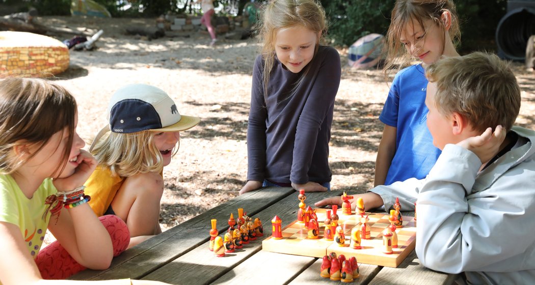 Fünf Kinder sitzen draußen um einen hölzernen Picknicktisch herum und spielen eine Partie Schach auf einem bunten Brett; im Hintergrund sind Bäume und Spielgeräte zu sehen.