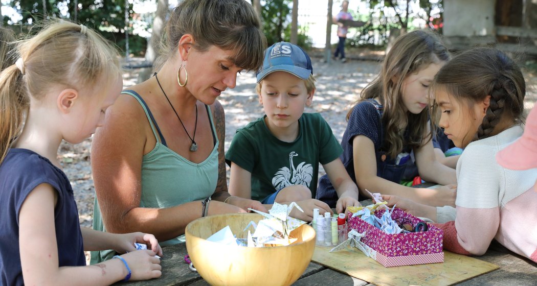 Eine Frau und fünf Kinder sitzen an einem Tisch im Freien und beschäftigen sich mit Bastelarbeiten. Die Bastelmaterialien sind ausgebreitet, und sie wirken konzentriert und arbeiten gut zusammen, während im Hintergrund Bäume zu sehen sind.