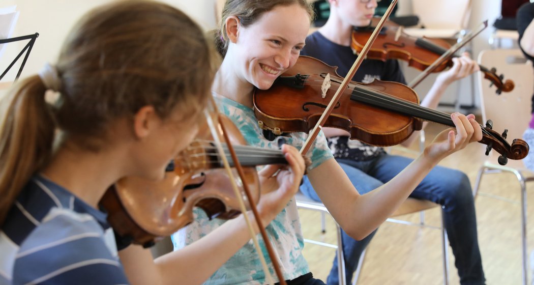 Drei junge Leute sitzen in einem Raum und spielen gemeinsam Geige. Die Person in der Mitte lächelt jemanden zu ihrer Linken strahlend an, und die Gruppe scheint Spaß an einer lebhaften Musikprobe zu haben.