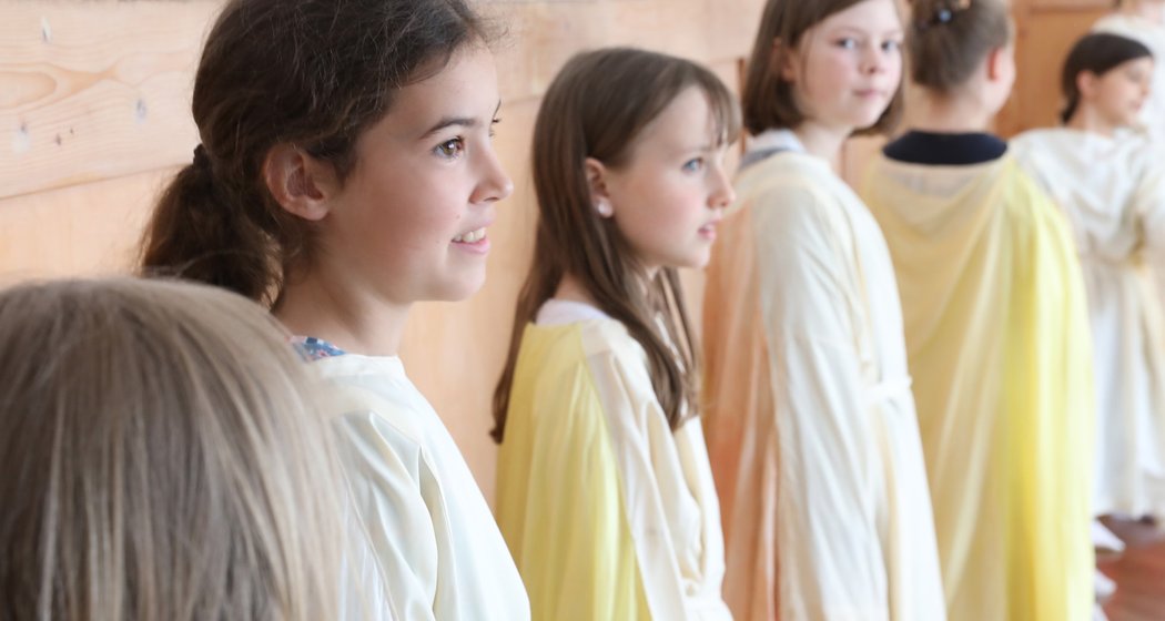 Mehrere kleine Kinder, überwiegend Mädchen, stehen in einem Raum in hellen Gewändern in einer Reihe. Sie blicken nach vorne, und die Szene wirkt hell und fröhlich. Im Hintergrund ist eine Holzwand zu sehen.