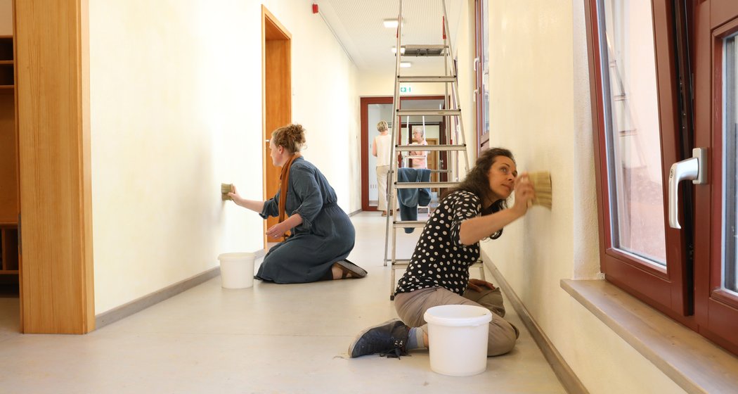 Zwei Frauen reinigen mit Schwämmen eine Wand im Flur, jede steht neben einem Eimer. Weiter hinten im Flur steht eine Person auf einer Leiter und streckt die Arme zur Decke. Durch die großen Fenster auf der rechten Seite fällt Sonnenlicht herein.