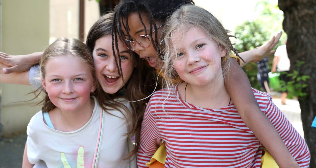 Vier lächelnde Kinder posieren eng beieinander im Freien; ein Kind trägt ein anderes auf dem Rücken, während sich die anderen umarmen – alle wirken im Tageslicht fröhlich und ausgelassen.