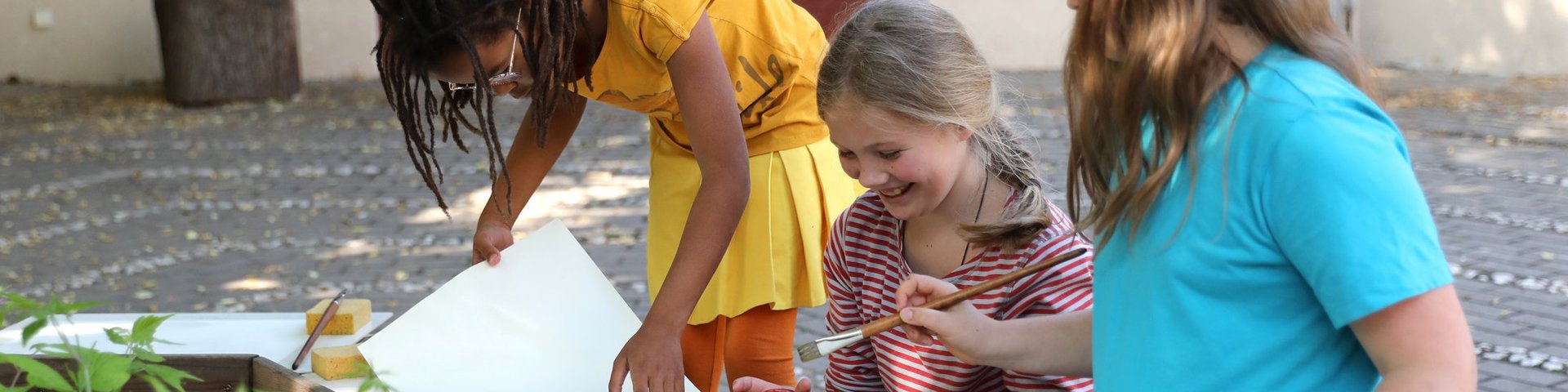 Drei Kinder sitzen draußen und bereiten sich darauf vor, mit Töpfen voller bunter Farben, Pinseln, Papier und Wasser auf einem Holztisch zu malen. Sie wirken glücklich und sind ganz in ihre kreative Tätigkeit vertieft.