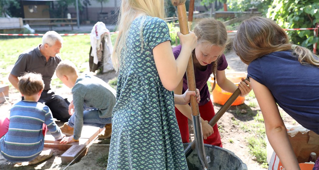 Die Kinder arbeiten im Freien; drei von ihnen mischen mit Schaufeln etwas in einer Wanne, während andere gemeinsam mit einem Erwachsenen Ziegelsteine stapeln. Sie sind von Grün und Sonnenlicht umgeben, was eine gemeinschaftliche, praxisorientierte Lernumgebung schafft.